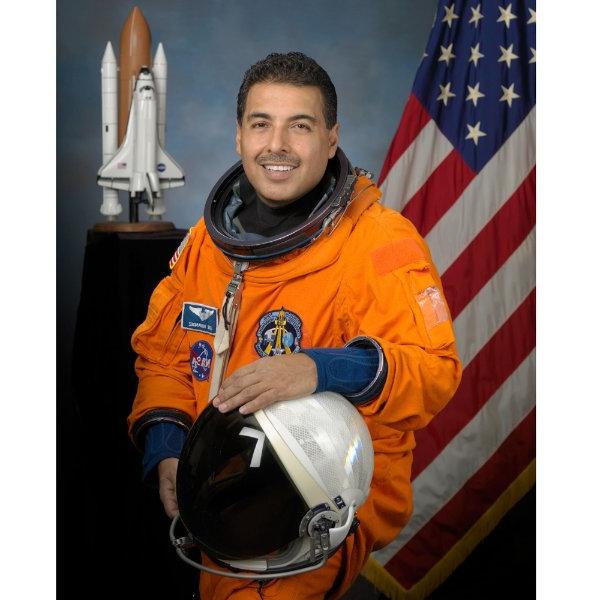 一个穿着橙色连体衣、戴着宇航员头盔的人坐在那里微笑. 背景是一面美国国旗和一枚火箭.