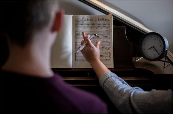 一个学生的背部和一个钢琴老师的手臂, pen in hand, gesturing toward a music book.