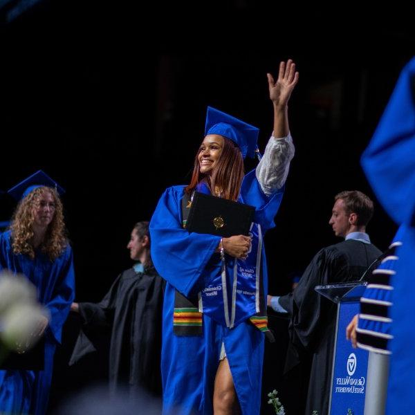 A person in a cap and gown walks across the stage with their hand up waving at the crowd.