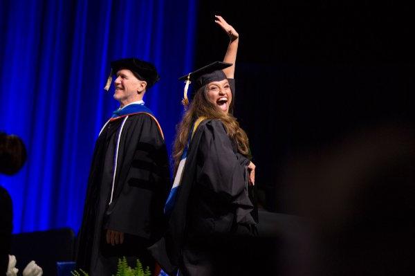一个头戴帽子、身穿长袍的人站在舞台上庆祝，一只手高举在空中，另一只手紧握着他们的文凭.