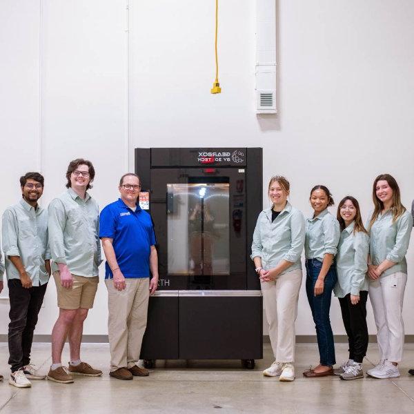 seven people, most in light green button down shirts, stand by a GearBox 3D printer in an industrial room in the Shape Corp. innovation design center